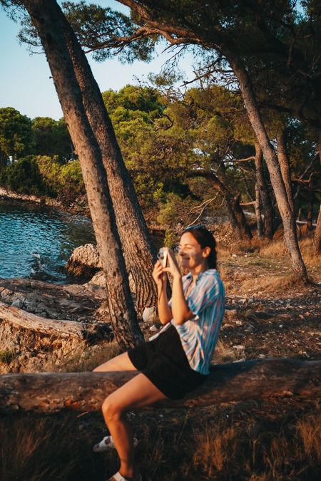 A young woman sitting on a log photographing the sea in Sibenik